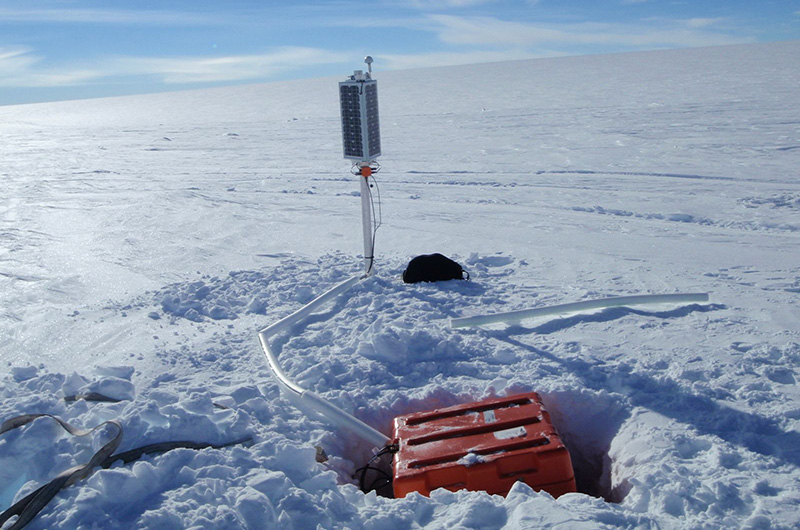 A sensor in an orange box buried in the vast Antarctica snow with a solar sensor on a post next to it